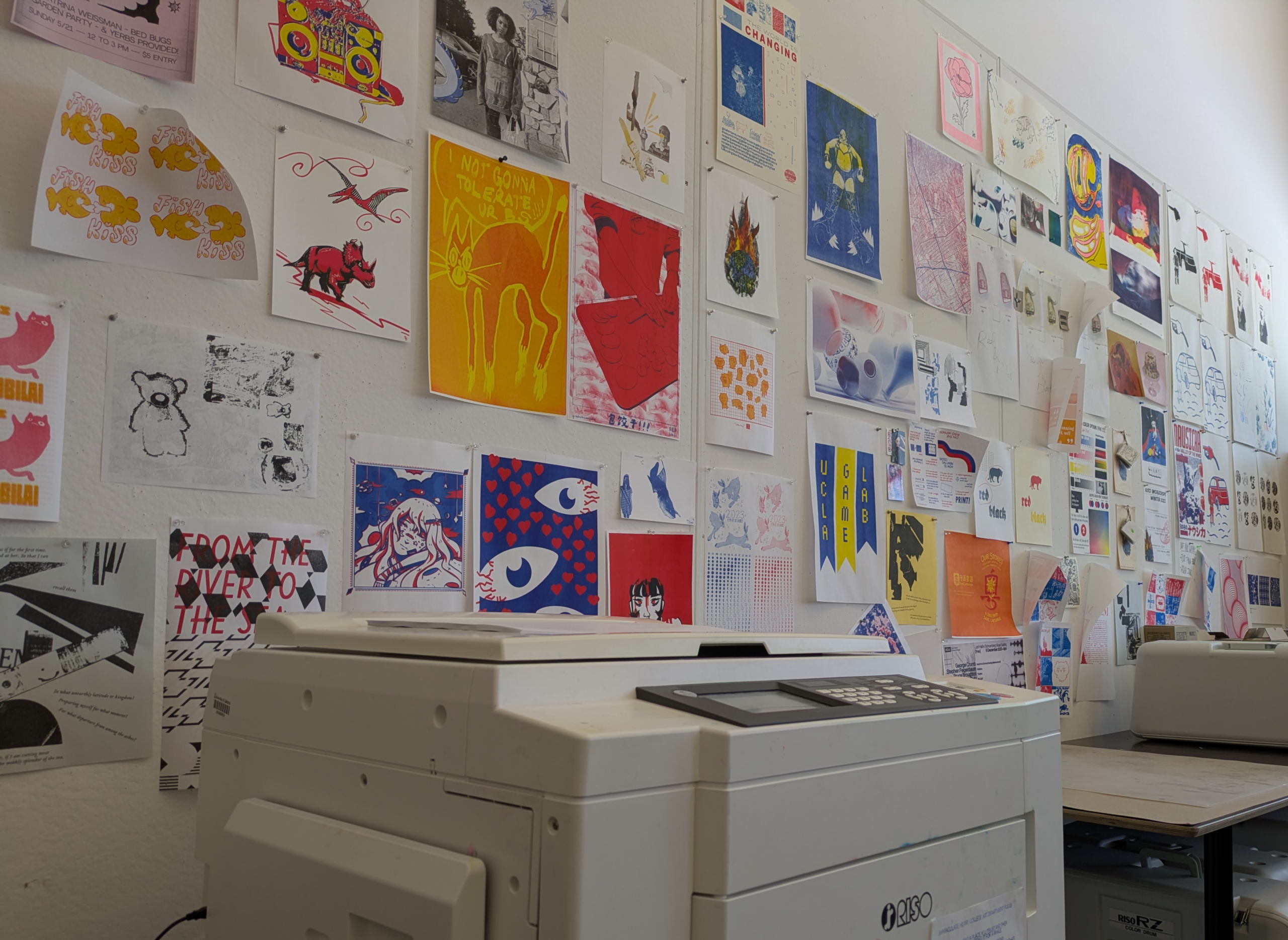 A Riso digital printer in the foreground in front of a wall of colorful prints of various sizes.