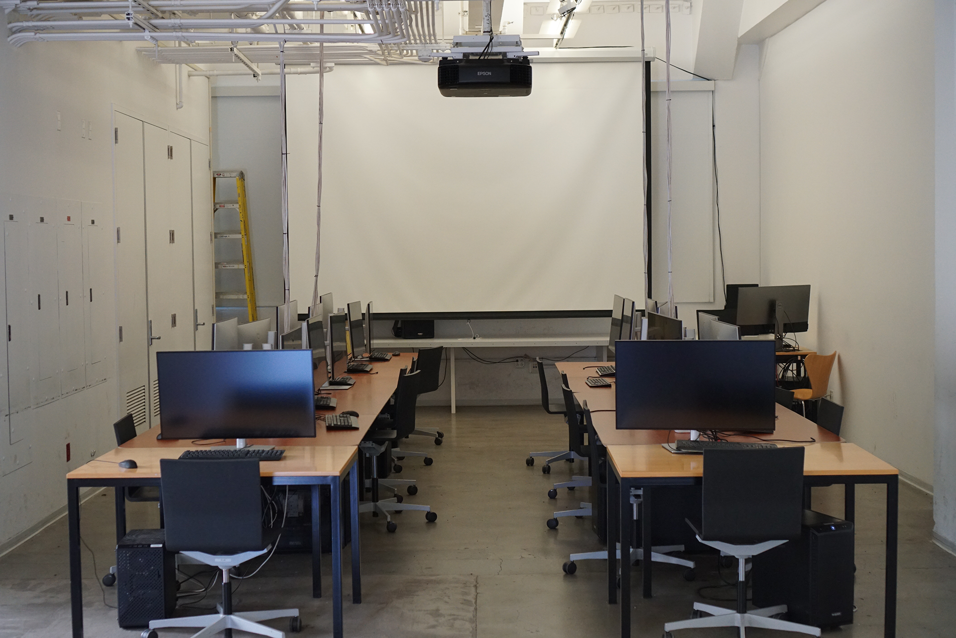 A bright computer lab known as the Last Classroom, featuring two parallel rows of wooden desks equipped with computer monitors and black rolling chairs, all facing a large, lowered projector screen at the front of a white-walled room.