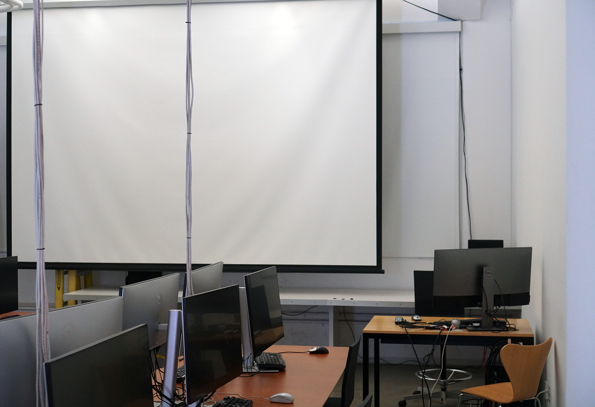 A close-up view of the front right section of the Last Classroom computer lab, focusing on a large, lowered projector screen. In the foreground, computer monitors line a wooden desk, while a separate, smaller workstation with a single monitor and a light wooden chair sits against the wall to the right. Cables can be seen hanging from the ceiling in front of the screen.