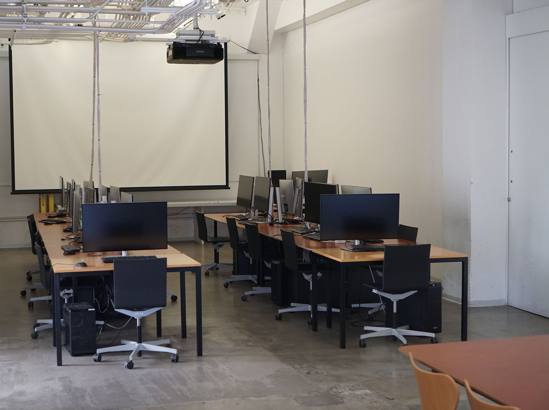 An angled view of the Last Classroom computer lab, showing rows of long wooden desks with computer monitors and black rolling chairs facing a large, lowered projector screen against a white wall.