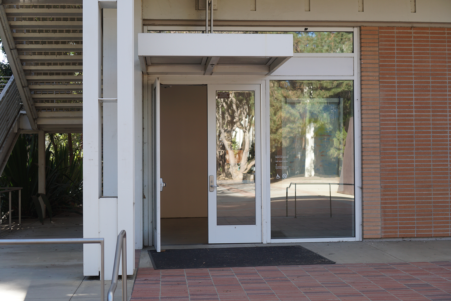 An exterior view of the New Wight Gallery entrance, featuring a white-framed double glass door with the left side propped open, situated beneath a small white awning between a metal staircase structure and a red brick wall.